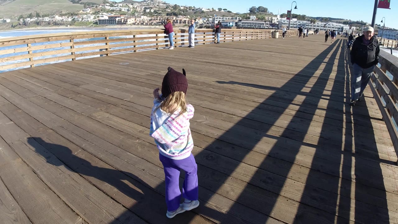 A girl in a purple hat and bright pants walks across the Pismo Beach pier, with a man near the right railing, people by the left rail, and the shoreline behind them.