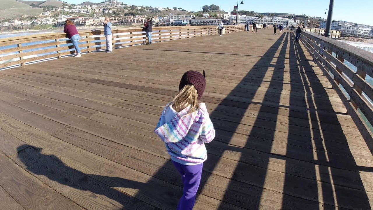 The blond child turns slightly to the right on the Pismo Beach pier, framed by diagonal railing shadows and a line of people in the background.