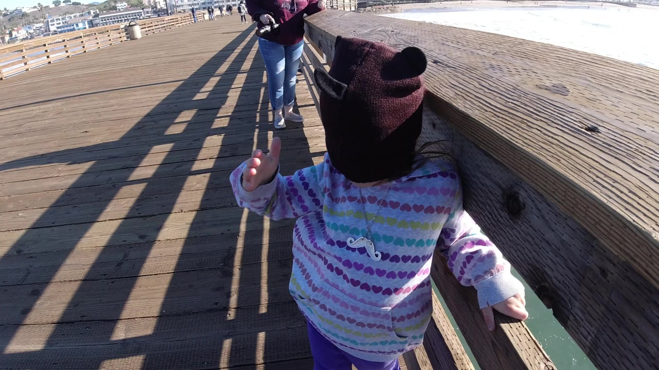 A young child in a dark knit cap and pastel hoodie stands beside a wooden railing on a Pismo Beach pier, with adults farther ahead and ocean water visible to the right.