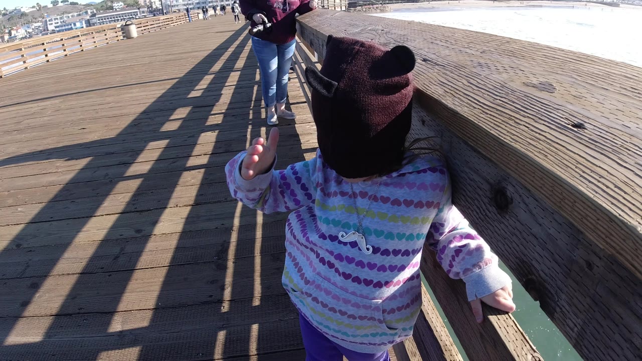 The child faces the wooden railing on the Pismo Beach pier with one hand lifted, while a woman stands ahead and bright water runs along the right edge.