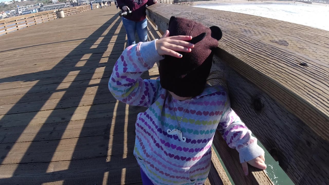 A young child lifts a hand across the dark knit cap while standing next to the wooden railing on the Pismo Beach pier, with sunlit ocean and pier boards visible around them.
