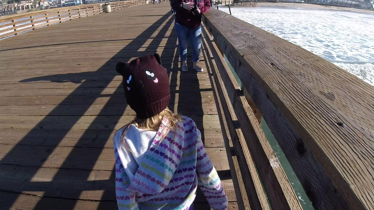 A young girl in a dark bear-eared knit hat and patterned hoodie walks ahead on a wooden pier at Pismo Beach, California, with surf visible beyond the railing.