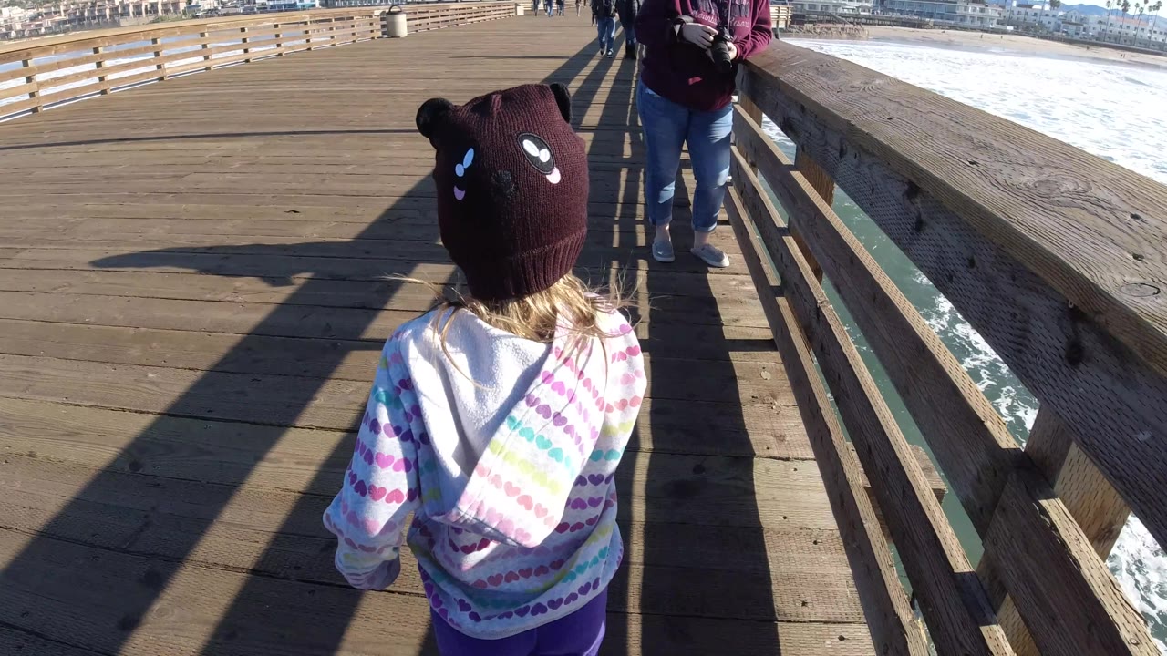 The child moves farther along the Pismo Beach pier toward an adult in jeans, while white surf runs alongside the railing to the right.