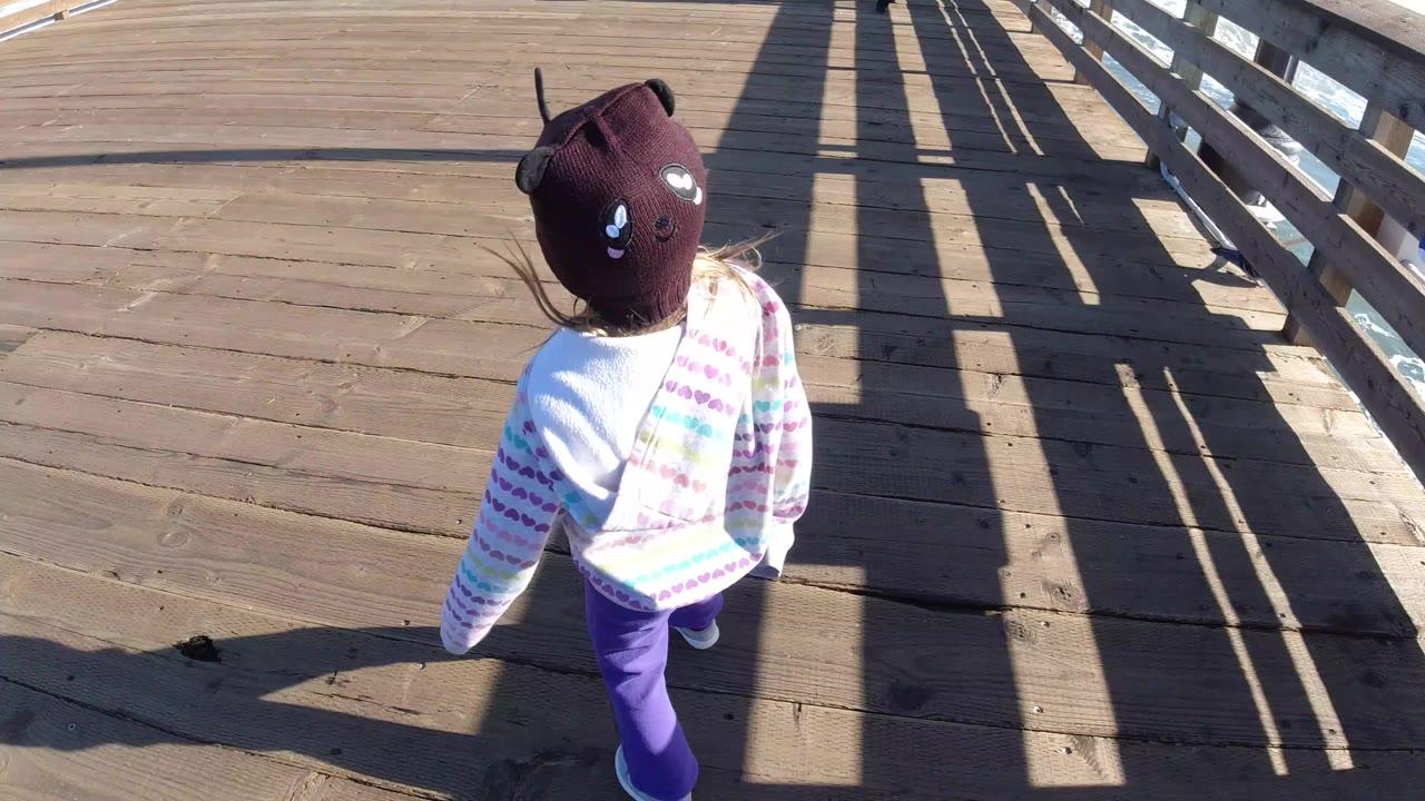 A child in a brown knit hat walks on a wooden boardwalk in Pismo Beach, California, seen from above with railings and long shadows across the planks.