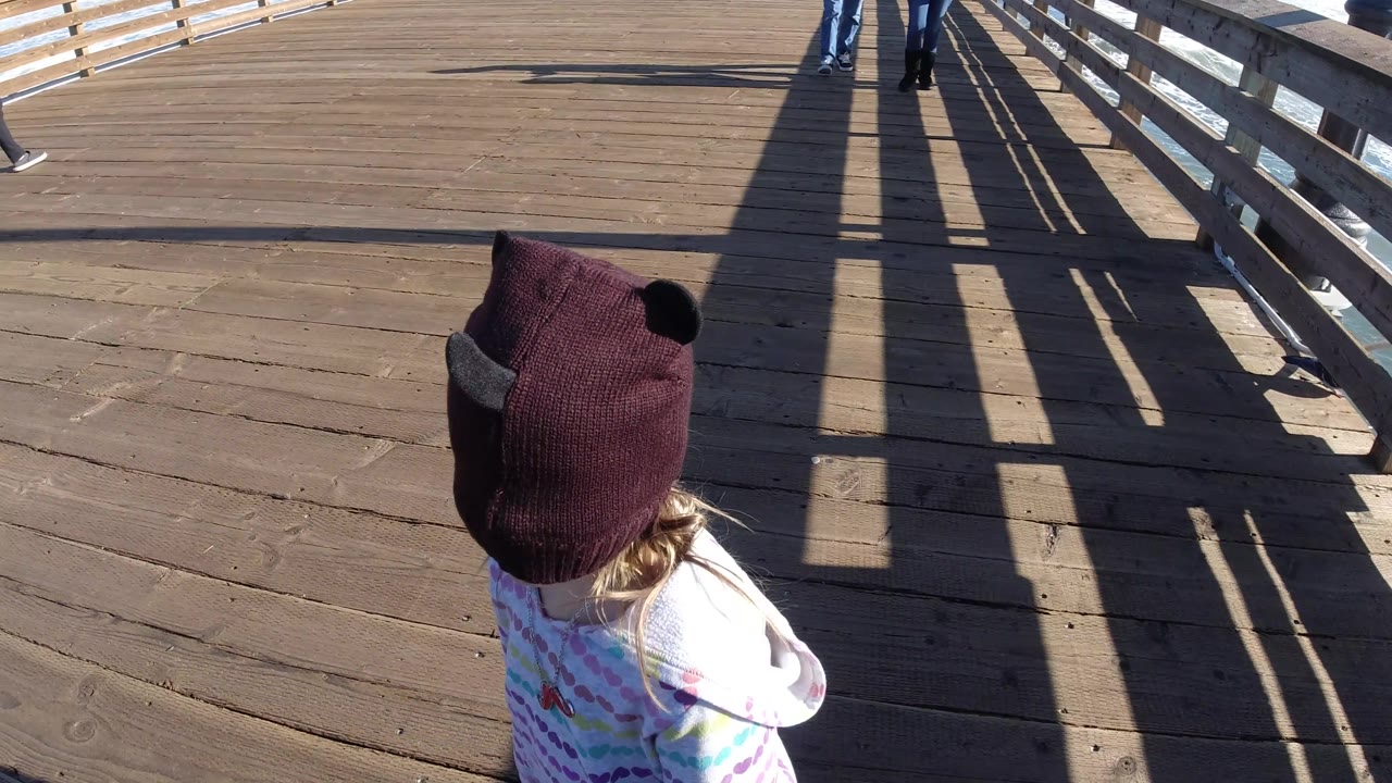 The child appears near the right edge of the Pismo Beach boardwalk frame while adult legs are partly visible ahead near the railings.