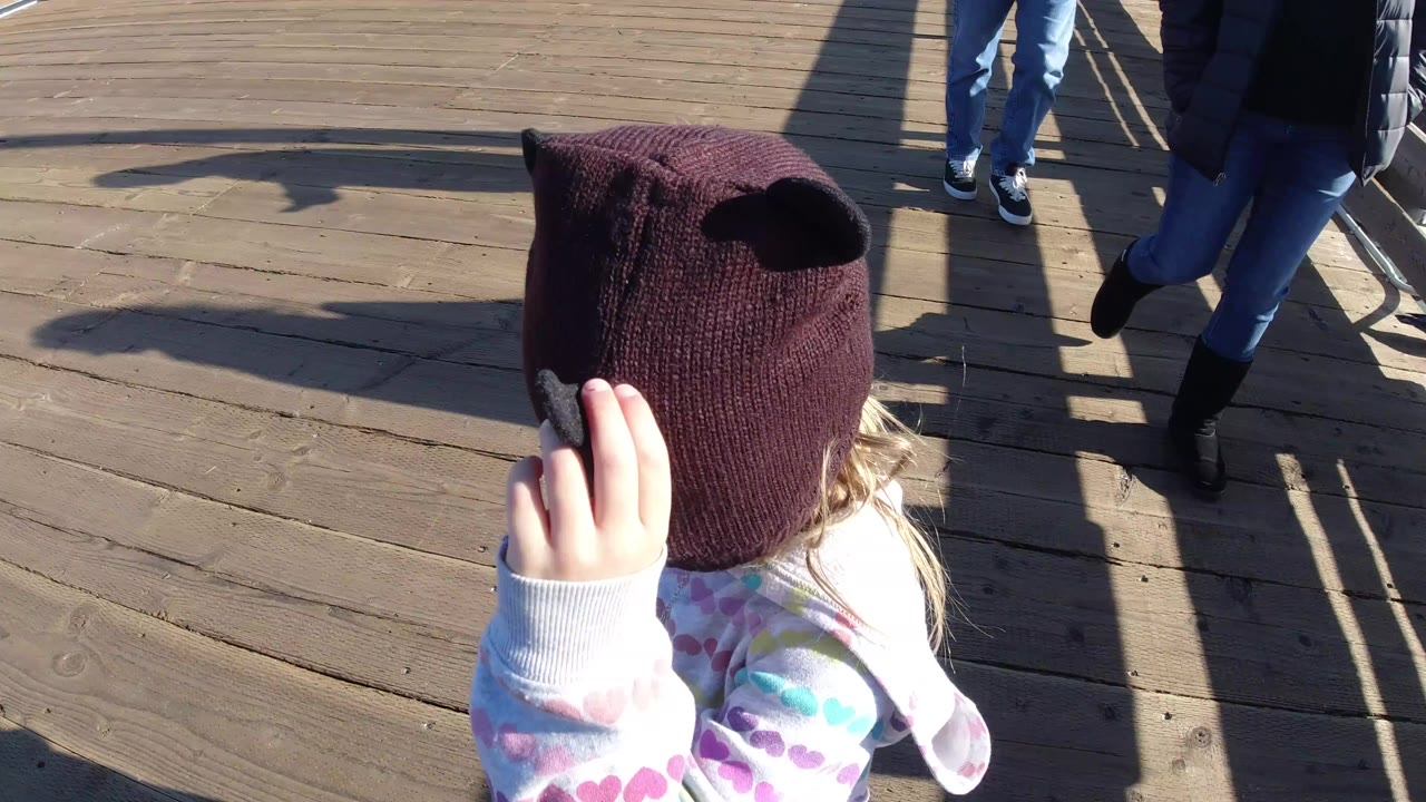 A young girl in Pismo Beach wears a dark brown knit hat and patterned sweatshirt on a bright wooden deck while adult legs appear in the background.