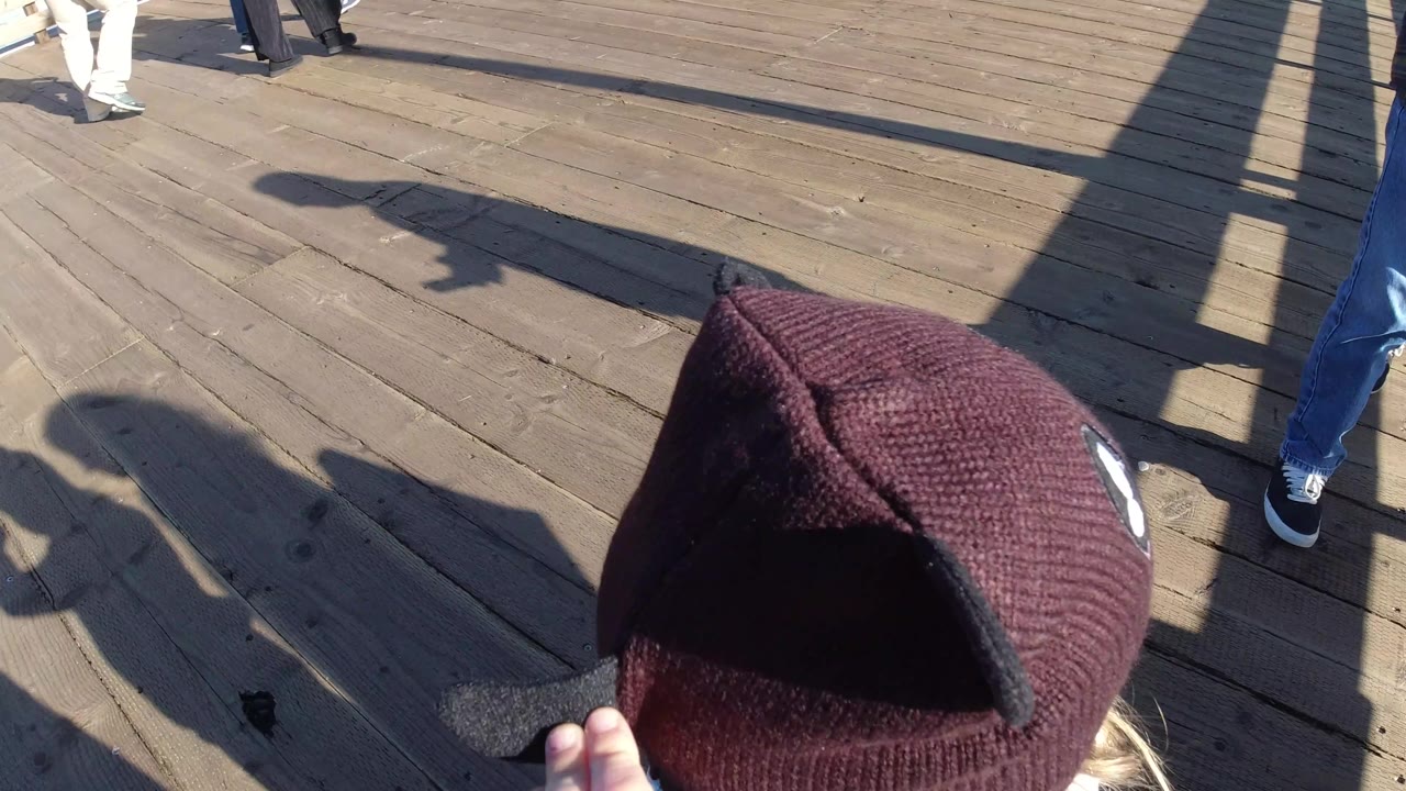 A close high-angle frame in Pismo Beach shows the child's dark brown hat and hand at the right edge, with adult feet and strong shadows across the deck.