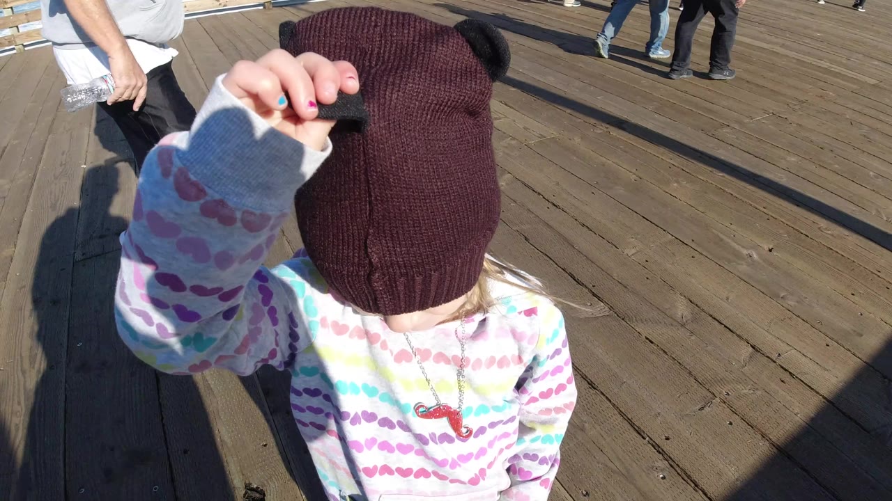 At Pismo Beach, a child in a dark knit beanie and pastel striped shirt stands close to the camera on wooden dock planks while adult legs pass nearby.