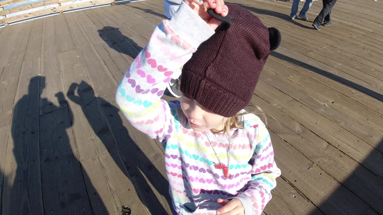 At Pismo Beach, the child turns toward the right side of frame with one arm up, while rail shadows and a few pedestrian legs appear across the wooden dock.