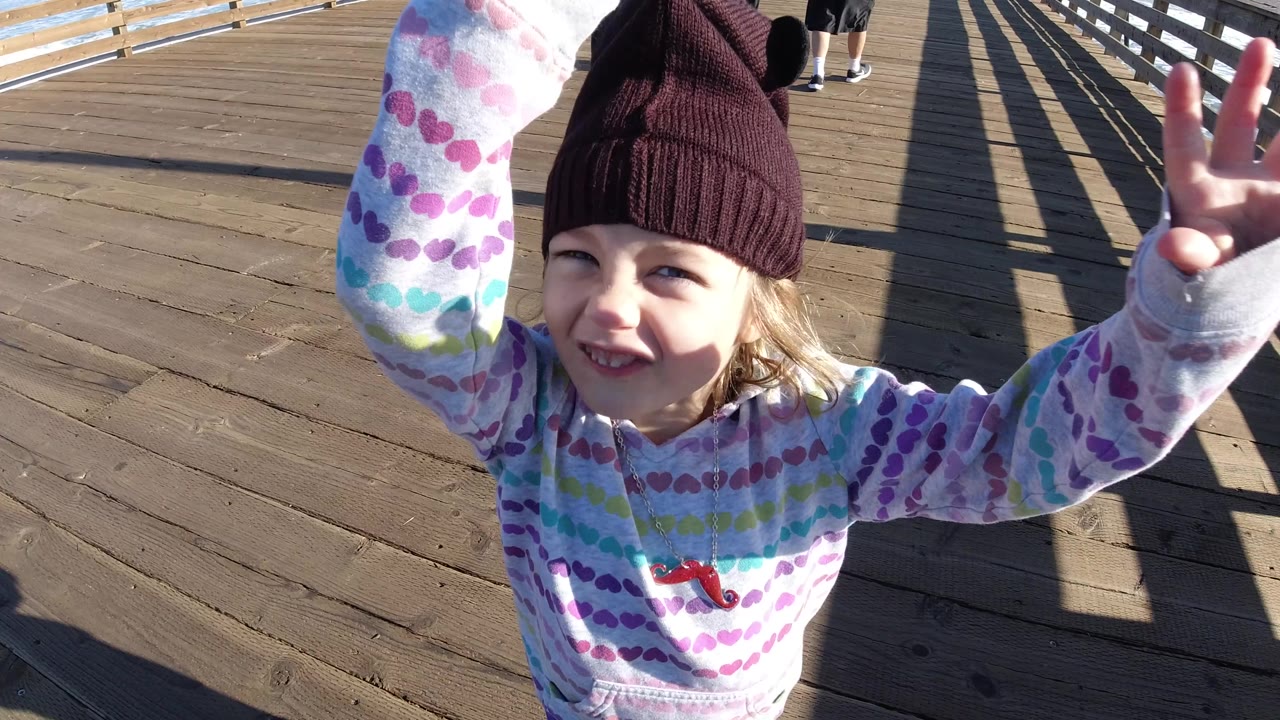 Young girl in a patterned hoodie smiles near the camera on a sunny Pismo Beach boardwalk while raising both hands, with railings and long shadows behind her.