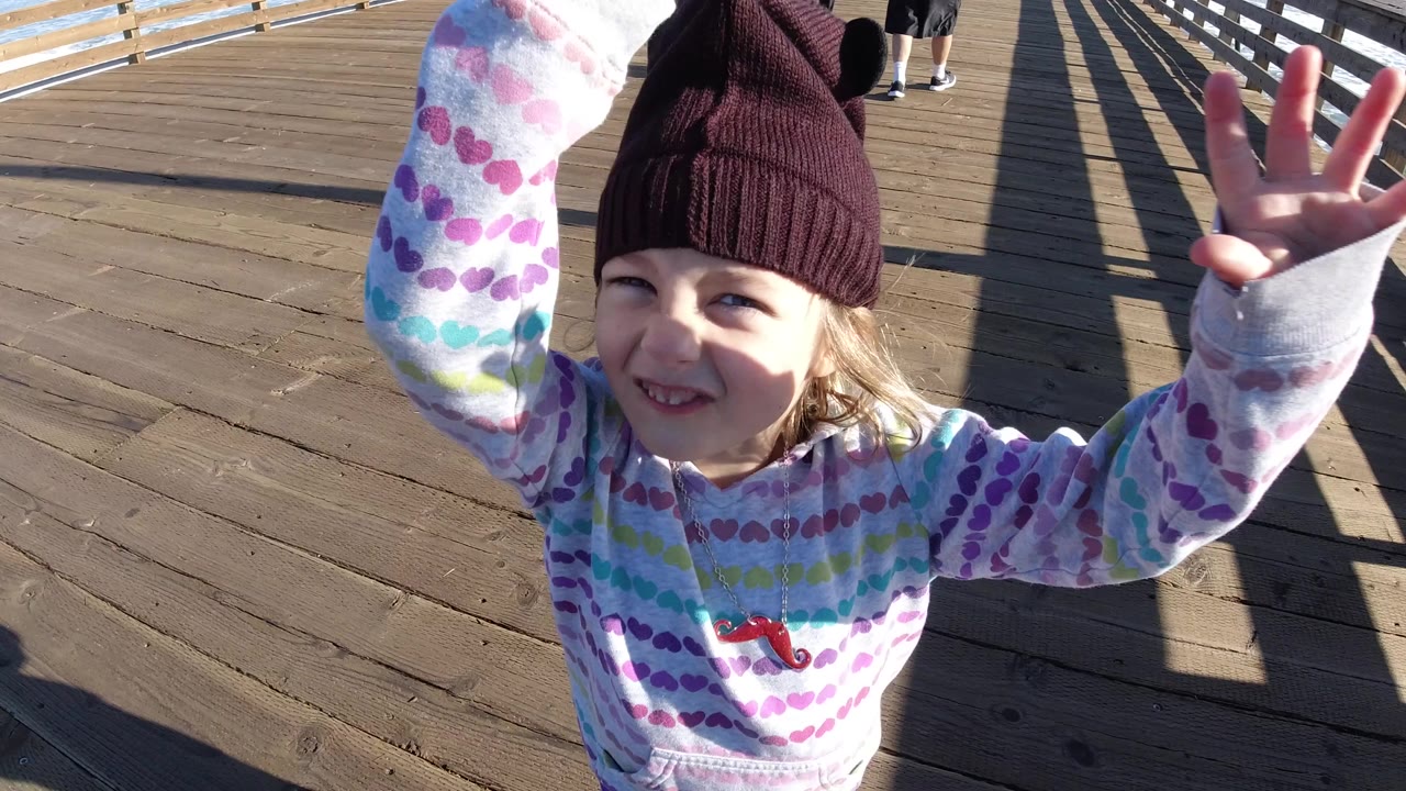 Young girl smiling with both arms lifted on a sunny wooden deck.