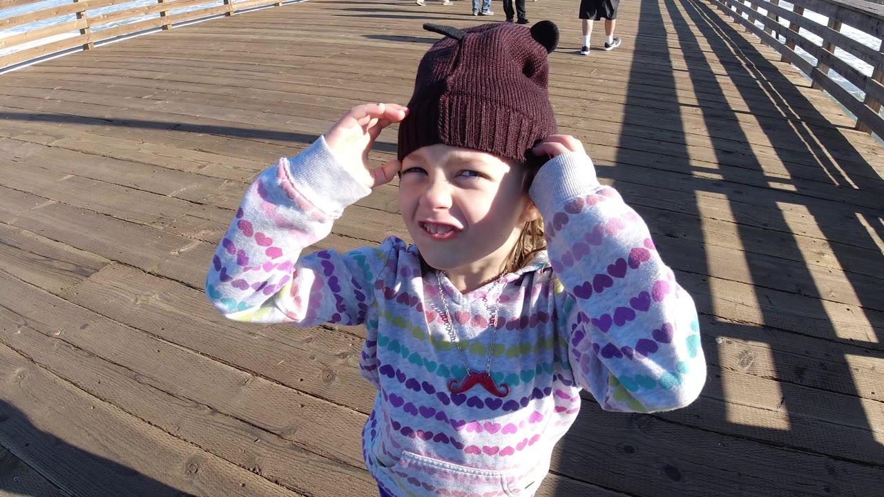 Young girl touches the sides of her brown knit hat on the Pismo Beach boardwalk, with the sun lighting her face and the railing shadows stretching behind her.