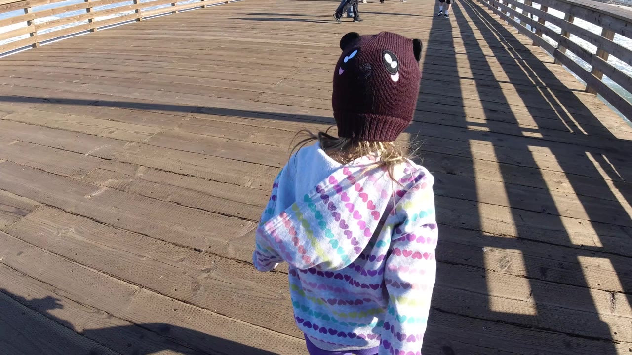 The child continues away from camera on the Pismo Beach pier, with the ocean beyond the right railing and long diagonal shadows stretching across the deck.