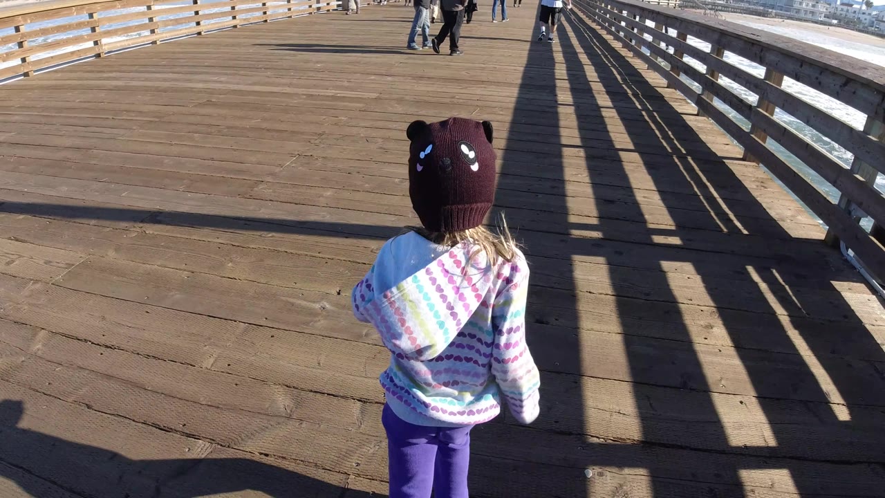 The young girl is smaller in frame on the Pismo Beach wooden pier, with surf visible through the right side and pedestrians walking farther ahead.
