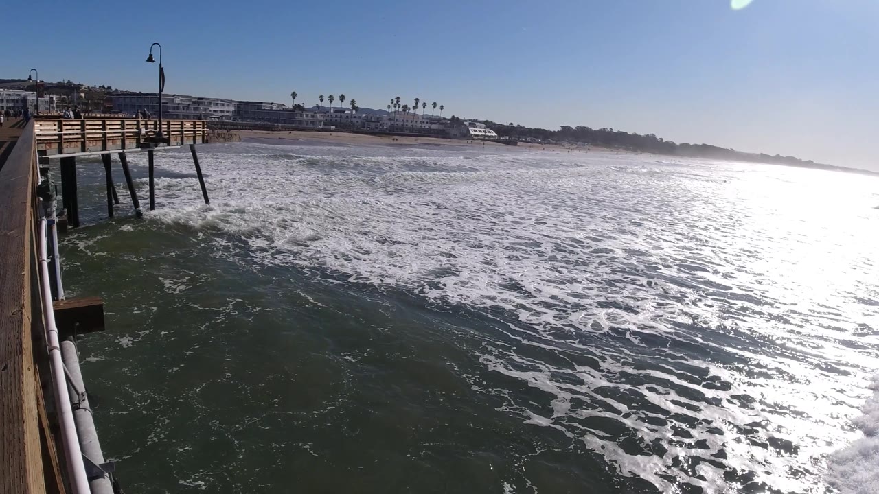 Pismo Beach boardwalk decking and vertical pilings appear at left while foamy waves fill the midground and bright glare washes the right side of the ocean, with no visible people.