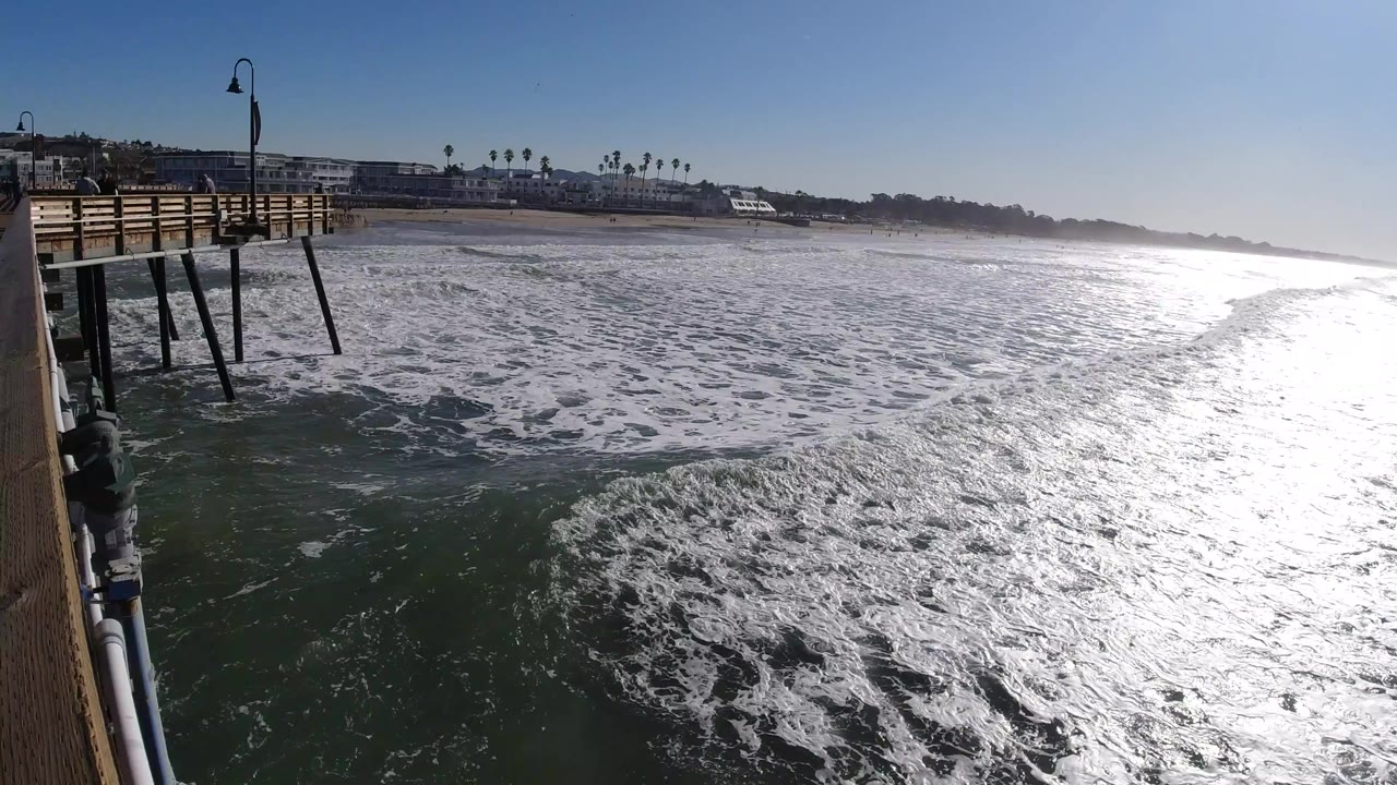 Opening frame over Pismo Beach water with the pier structure at left, foamy surf across the center, bright reflection on the right side of the ocean, and no visible people.