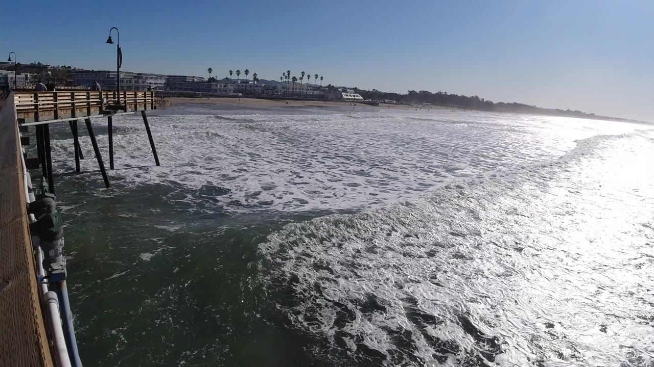 Second frame at Pismo Beach shows the wooden pier and lamp post on the left, broad whitewater below, distant beachfront buildings and palms on the horizon, and no visible people.