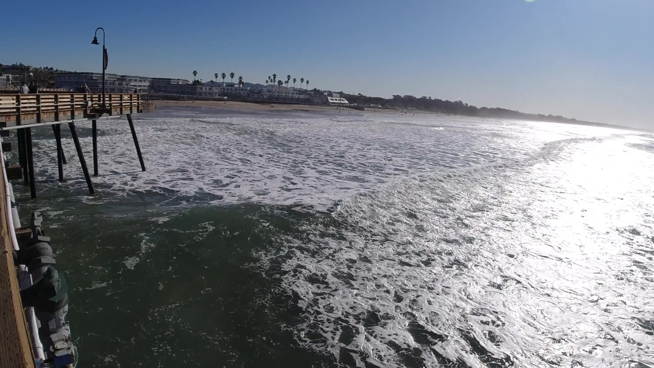 At 0:02, the Pismo Beach pier pilings extend down into darker water at left while dense white foam and a wide band of sun glare cover the right side, with no visible people.