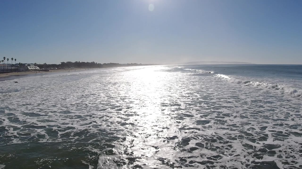 At Pismo Beach, California, the sun glare widens over wet shoreline water while low waves break to the right, with no visible people.