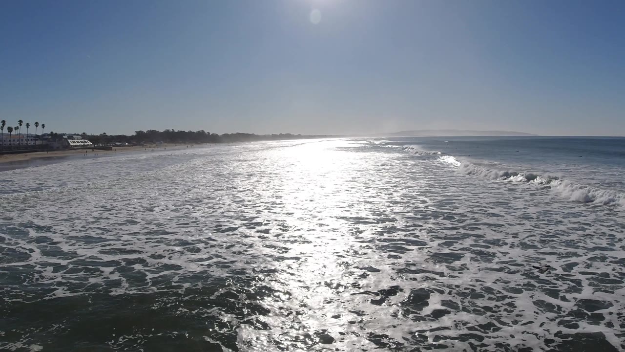 At Pismo Beach, California, dense white foam covers the foreground water while the open ocean extends right of the sunlit shoreline, with no visible people.