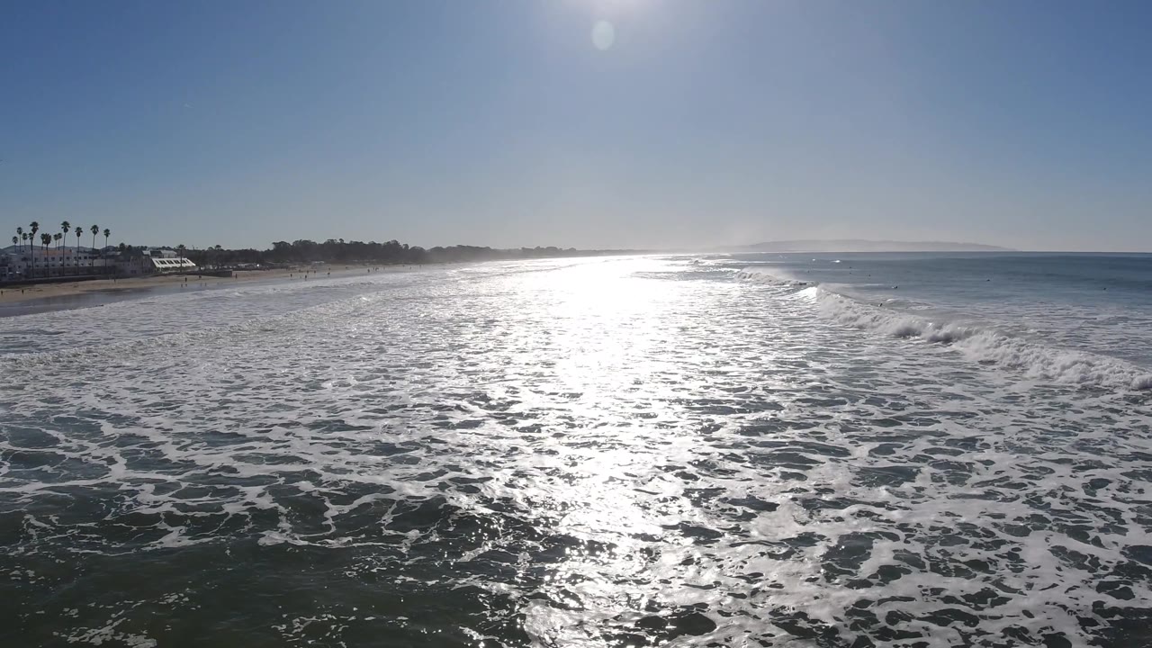 At Pismo Beach, California, a wide band of whitewater spreads across the beach as sun glare runs from the horizon toward the foreground, with no visible people.