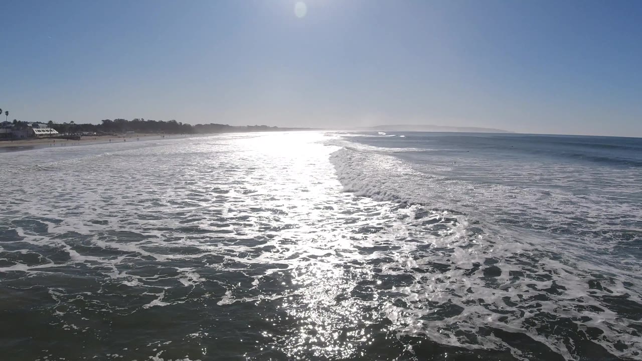 The Pismo Beach shoreline is more visible at left, with palm trees and low buildings beyond the surf while the sun reflection dominates the center water and no visible people appear.