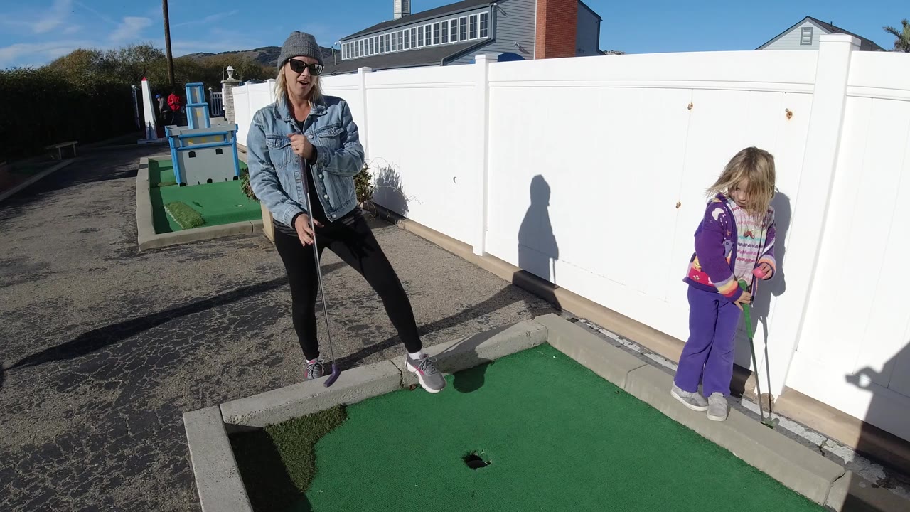 The woman stands back on the mini-golf lane while the child faces the white fence with a club, with the hole and sunlit turf visible in front.