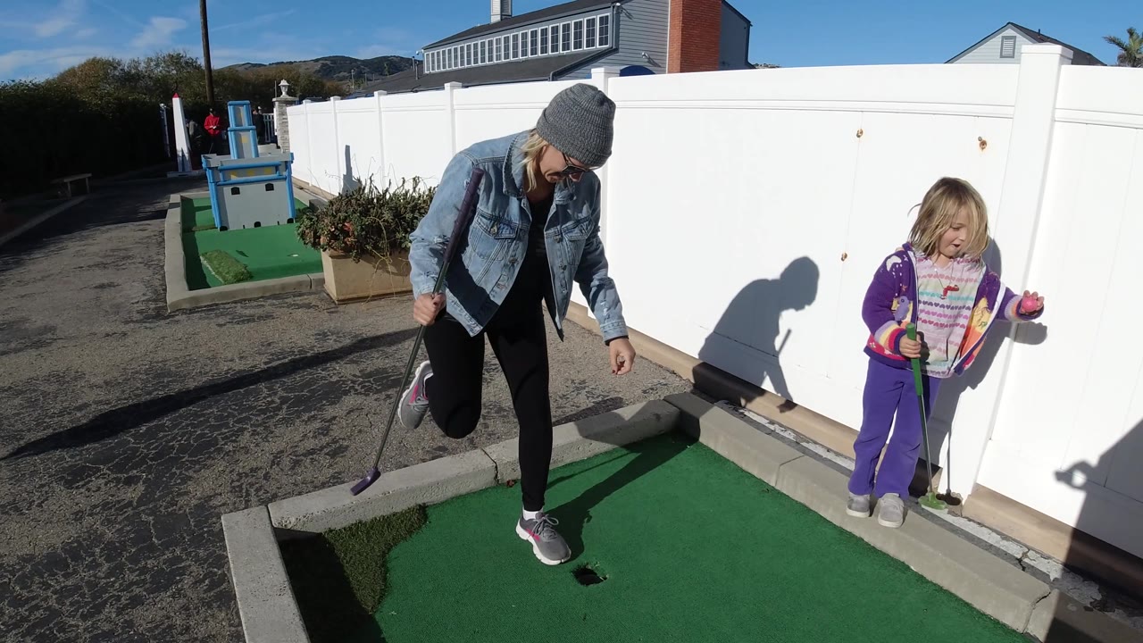 The woman steps across the green mini-golf turf near the hole as the child stands to the right with a club, both casting strong shadows on the fence and ground.