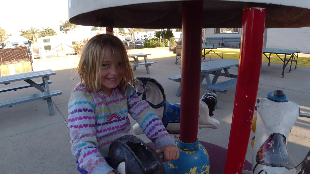 At Pismo Beach, California, a young girl faces the camera while riding a miniature merry-go-round horse beside red support poles and picnic tables.