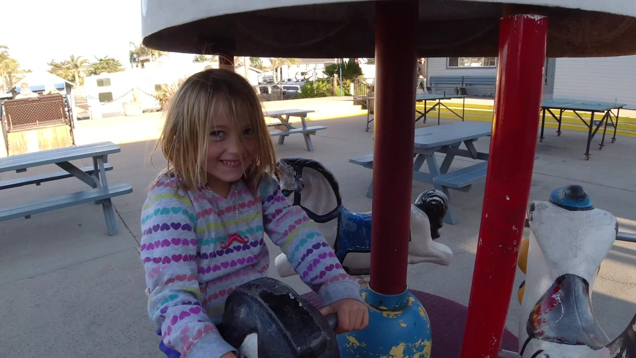 At Pismo Beach, California, the girl remains near the front of the frame on the small merry-go-round, with a white carousel horse and picnic tables visible behind her.