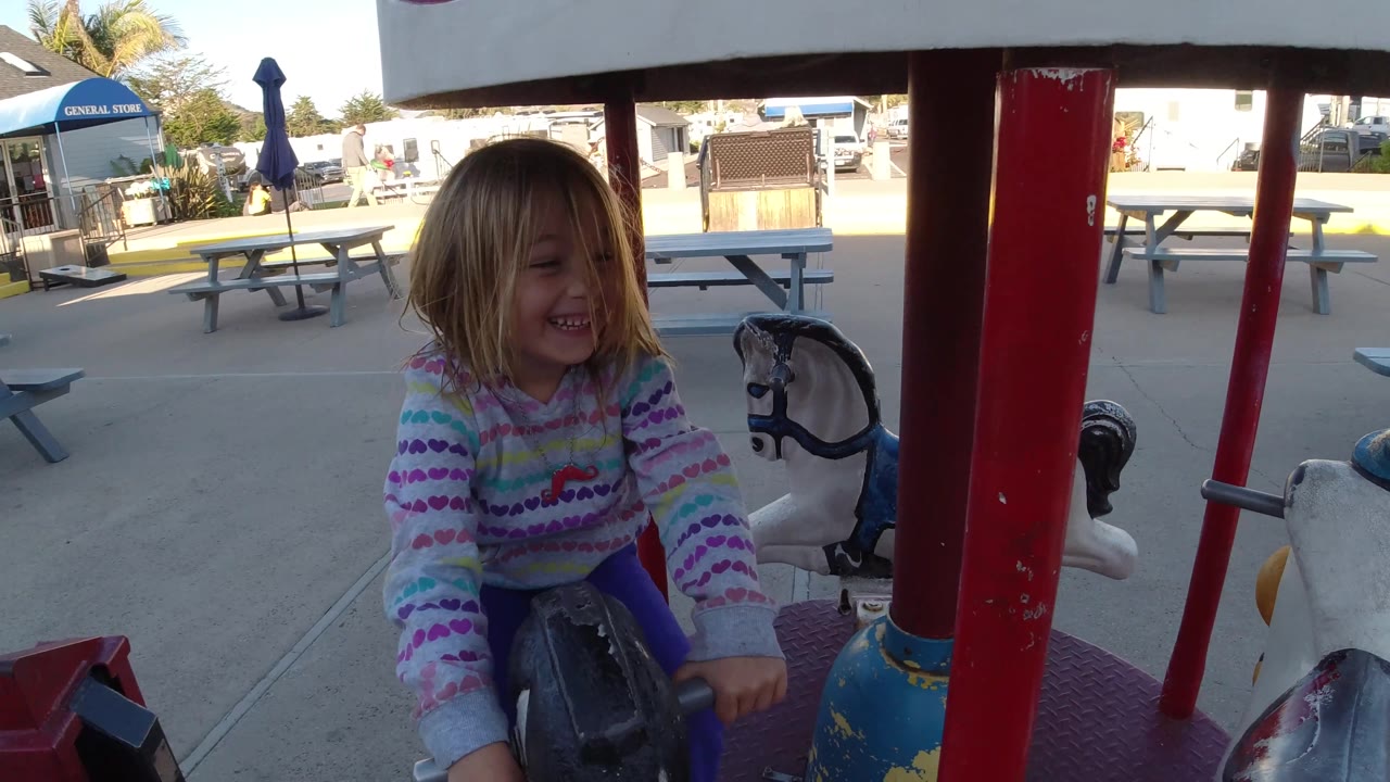 At Pismo Beach, California, the girl smiles while riding the small carousel horse, framed by red poles, a white ride canopy edge, and outdoor seating in the background.
