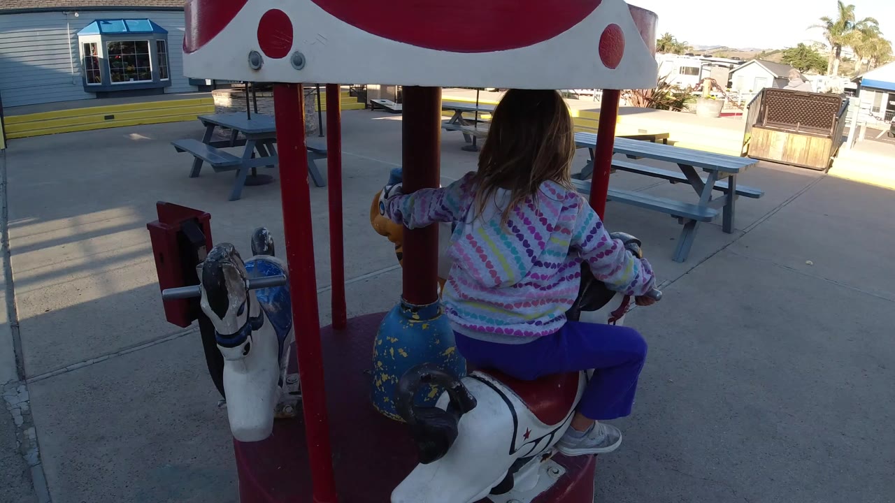 A toddler in a patterned top and purple pants sits on a painted animal seat of a playground carousel in Pismo Beach, with picnic tables and buildings behind the ride.
