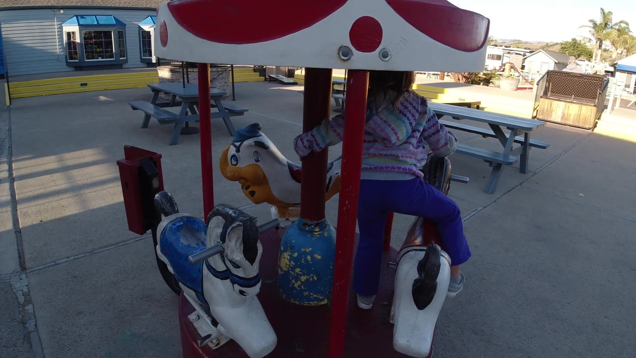 A toddler shifts position on the carousel in Pismo Beach, with a yellow painted animal face, white horse figure, red posts, and picnic tables visible across the paved play area.