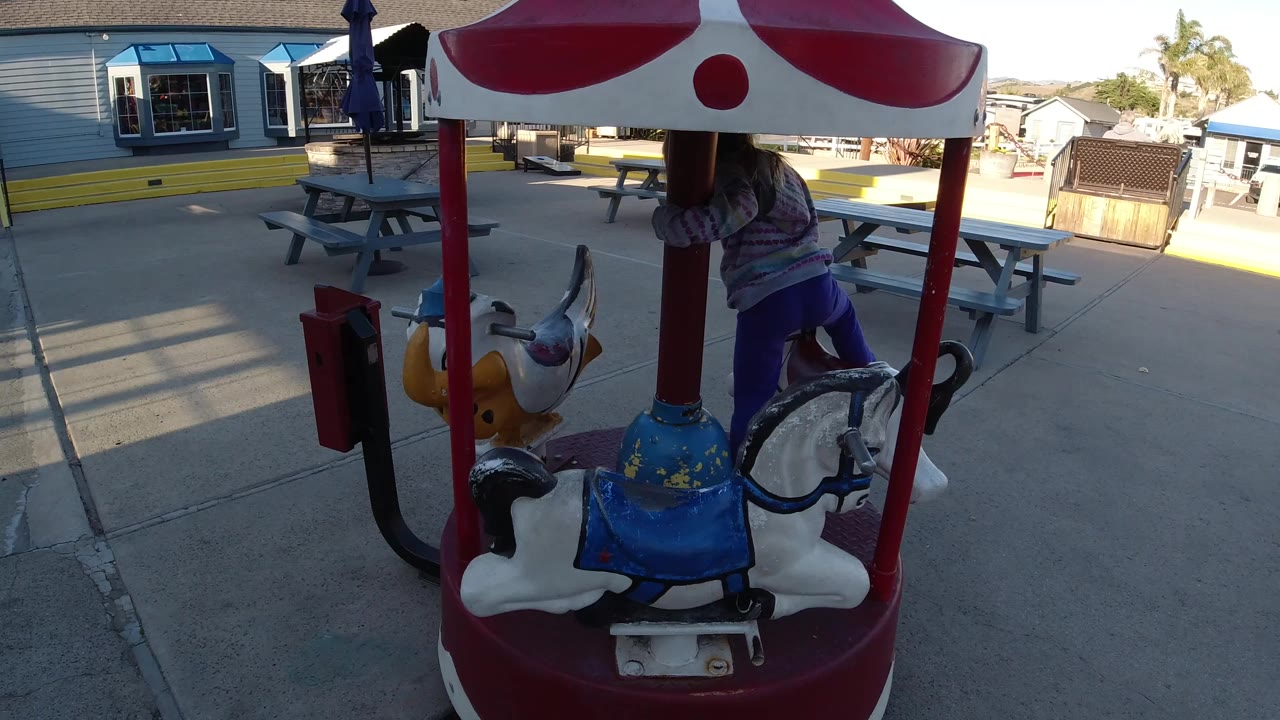 At 0:00 in Pismo Beach, California, a young blonde girl is partly obscured by the red and white canopy while moving around a small animal-seat merry-go-round.