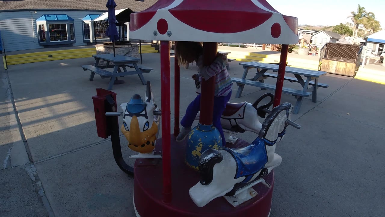 At 0:02 in Pismo Beach, California, the child leans near the front yellow animal seat as the carousel canopy and red posts frame the paved outdoor area.