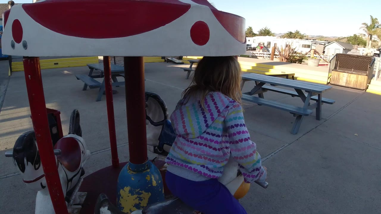 A young girl in a striped hoodie is seen from behind on a duck-shaped carousel ride in Pismo Beach, California, with picnic tables and low buildings beyond the ride canopy.