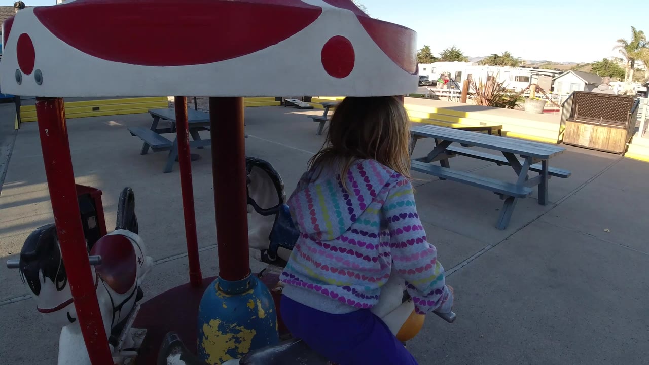 The girl remains seated under the red and white canopy of the small carousel ride in Pismo Beach, California, with the amusement area and benches visible ahead.