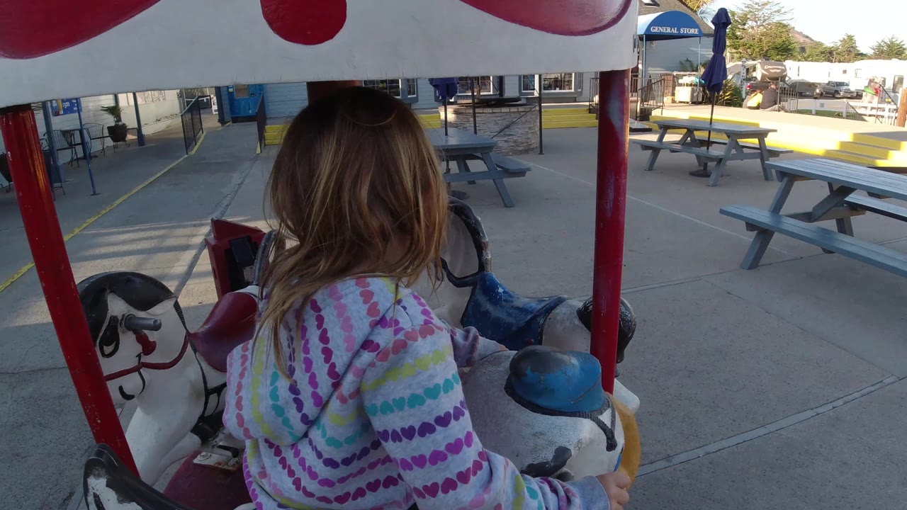 A closer side view shows the young girl on the ride with painted duck and horse figures, red poles, storefront windows, and picnic tables in Pismo Beach, California.