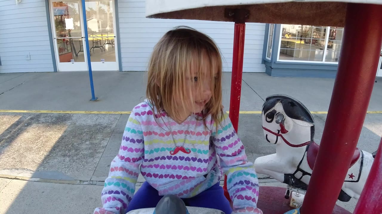 At 0:00 in Pismo Beach, California, a child in a rainbow hoodie sits close to the camera on a small ride while a white carousel horse and storefront windows sit behind them.