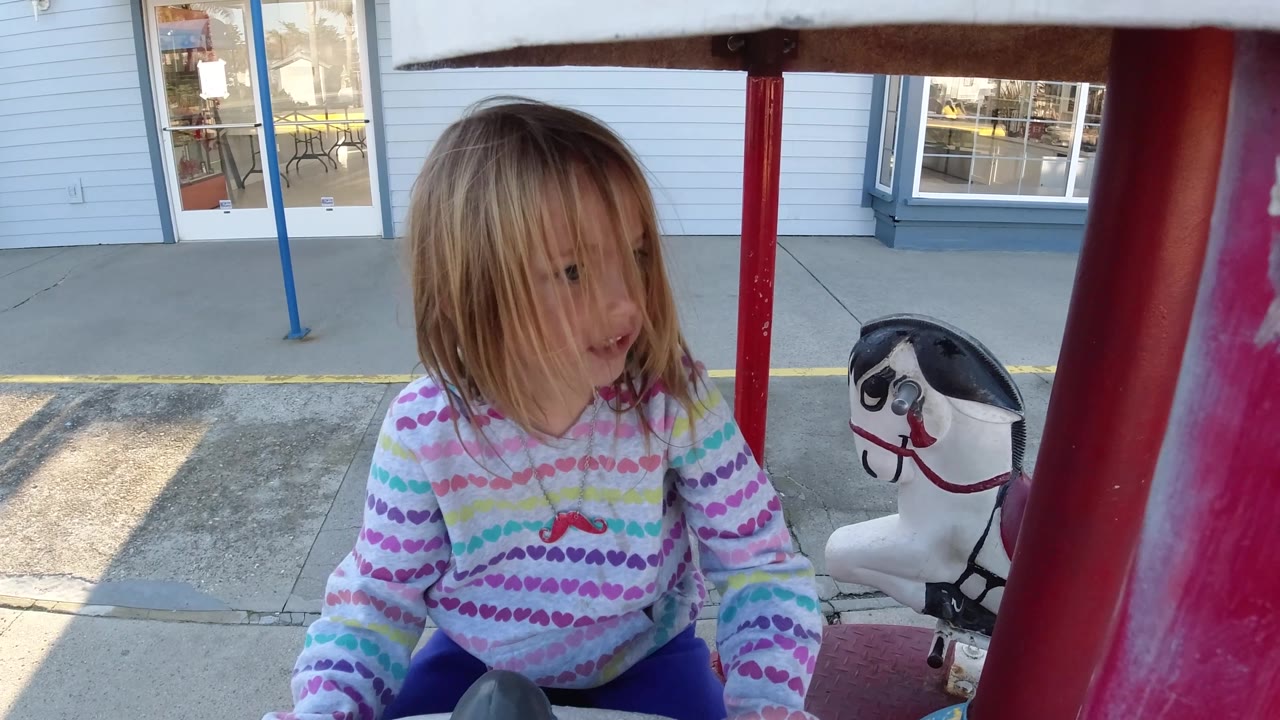 At 0:02 in Pismo Beach, California, the child looks slightly to the side while seated on the ride, with the red support pole and white carousel horse still visible behind them.