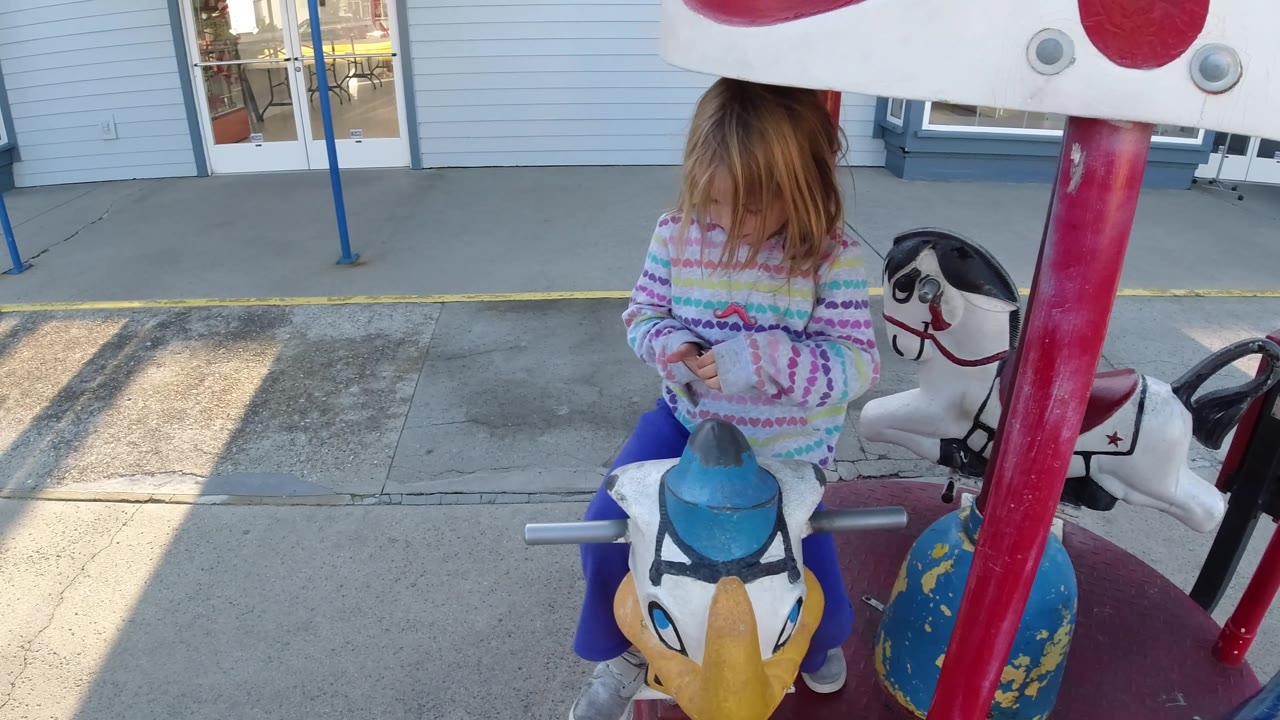 A young girl sits forward on the merry-go-round animal in Pismo Beach, partially framed by a red support pole with the white horse figure close behind her.