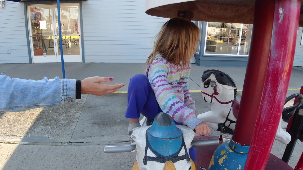 At the start of the Pismo Beach clip, a young girl sits on a white carousel horse while an adult hand reaches in from the left near the ride controls.