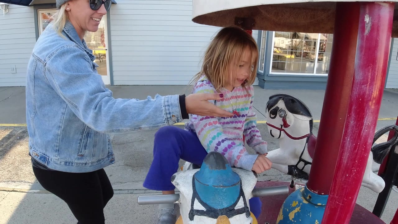 At 0:02, an adult woman in a denim jacket stands beside the seated child on the small carousel, with the ride poles and storefront facade behind them in Pismo Beach.