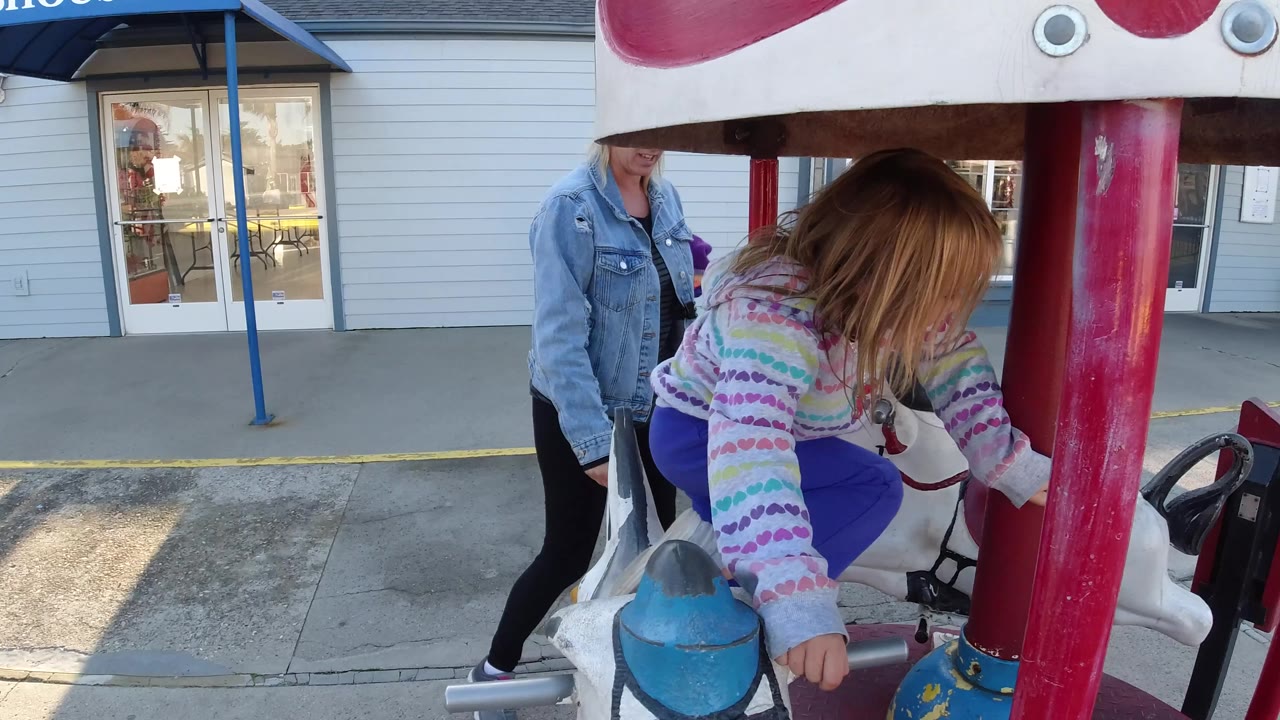 The Pismo Beach merry-go-round fills the foreground as the girl bends over the saddle and the adult remains just behind the ride frame.