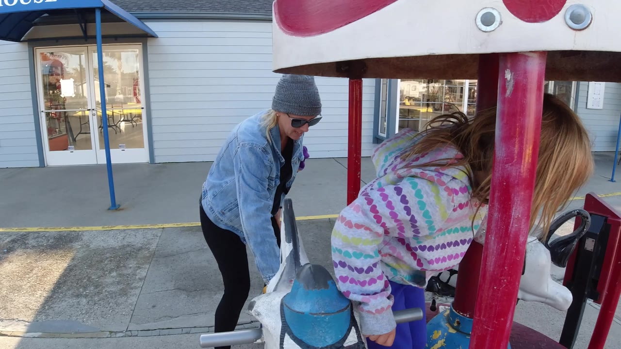 At the Pismo Beach ride, the adult in sunglasses and a gray beanie bends toward a painted carousel animal while the girl leans forward beside a red pole.