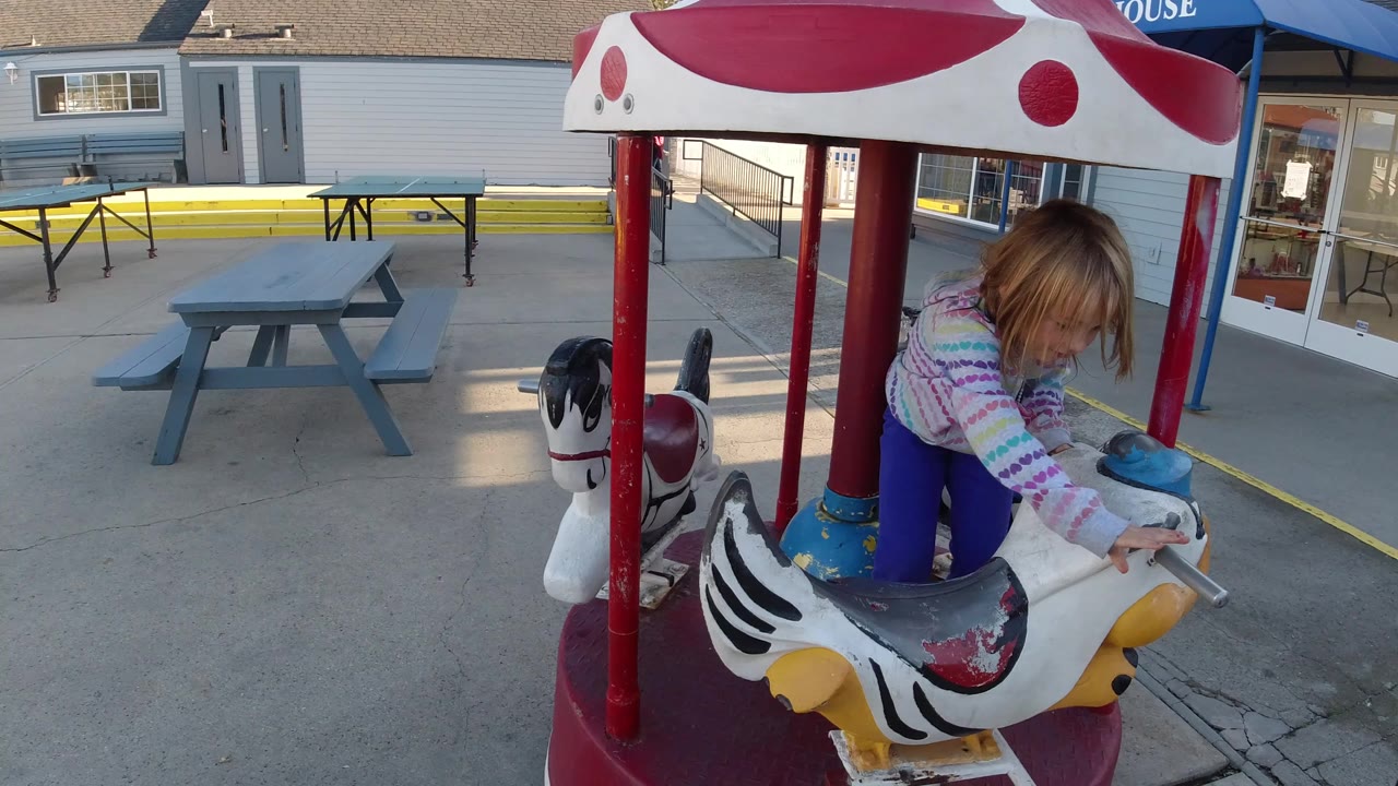 At 0:01 in Pismo Beach, the girl remains on the white carousel animal as red poles frame the ride and a picnic table sits across the concrete patio.