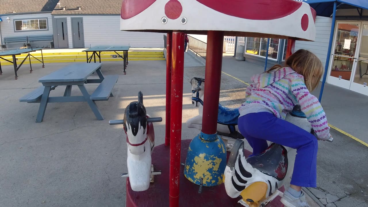 At 0:02 in Pismo Beach, the child is viewed from behind on the carousel, with white and blue animal figures, chipped paint, red poles, and storefront glass in the background.