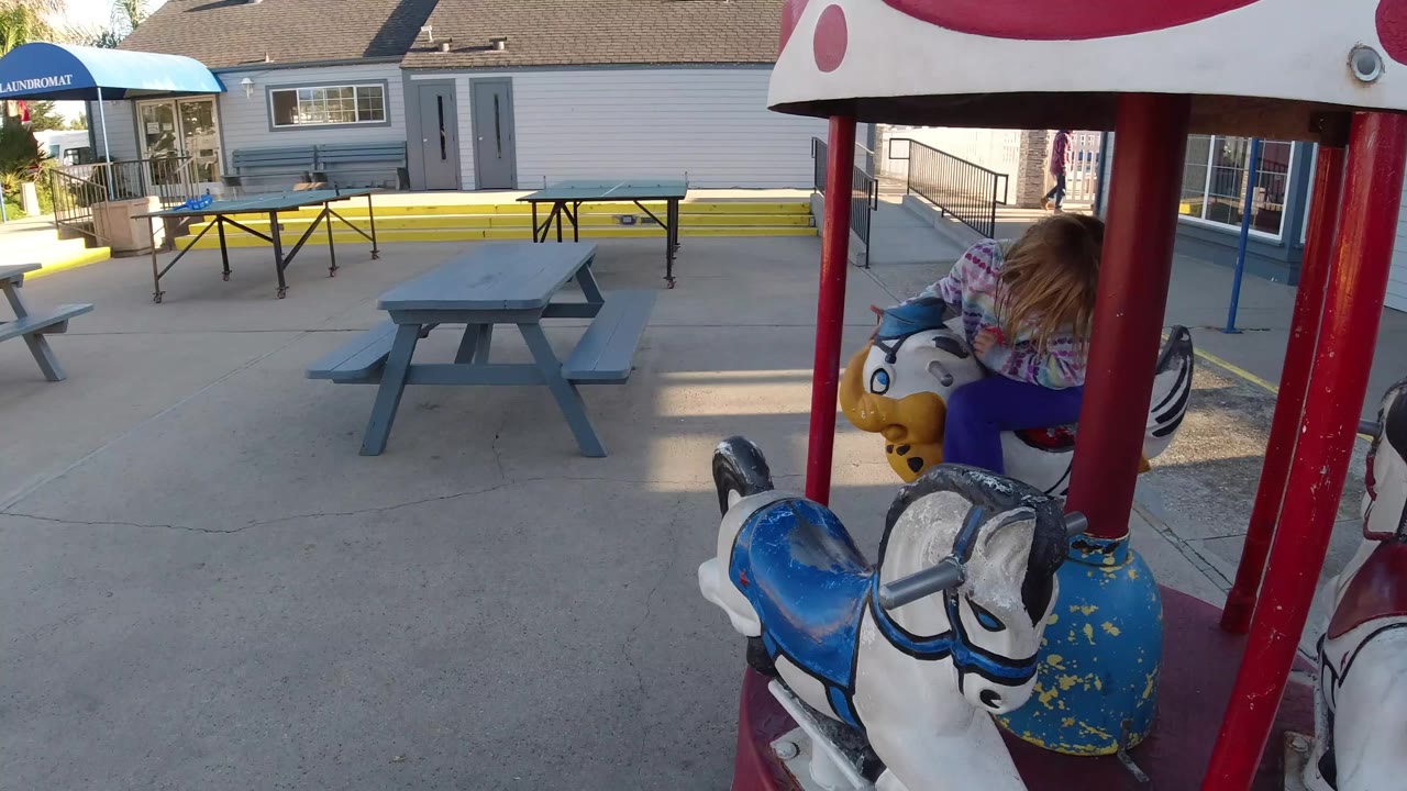 At 0:00 in Pismo Beach, California, a young blonde girl is partly obscured by red carousel poles while sitting on a horse-shaped ride in a concrete yard.