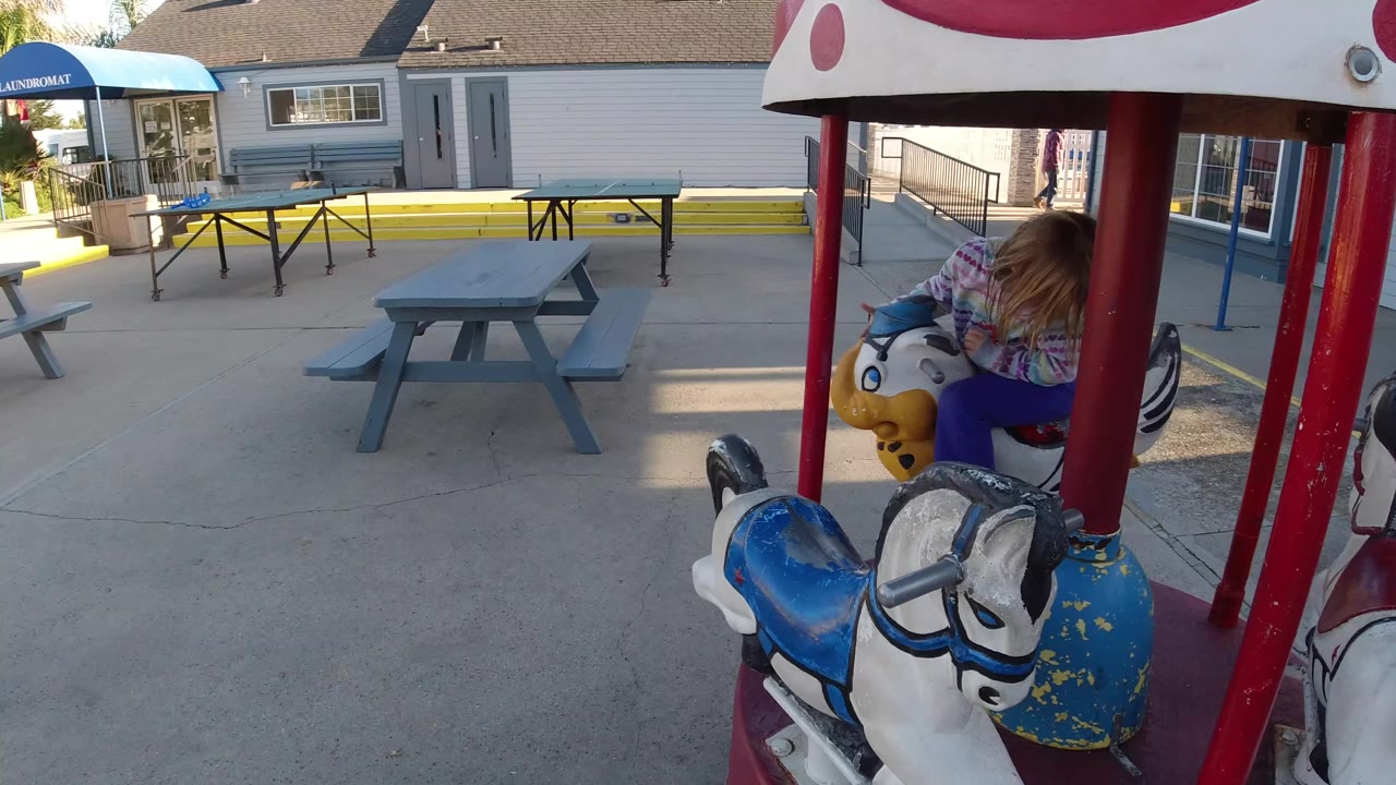 At 0:01 in Pismo Beach, California, the girl remains mostly turned away on the carousel, with a blue-and-white horse in the foreground and picnic tables across the yard.
