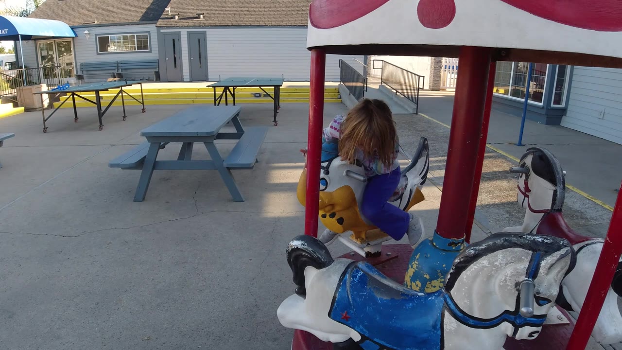 At 0:02 in Pismo Beach, California, the child is seen from behind on the carousel horse, with the red supports framing picnic tables, railings, and building doors in the background.